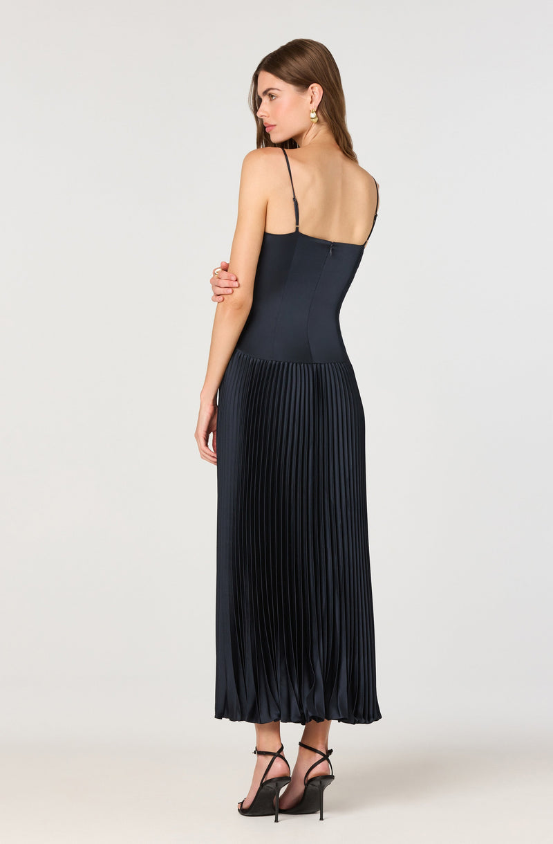 A woman is wearing a elegant, sleeveless, navy blue dress with thin spaghetti straps. The upper part of the dress is fitted and smooth, while the lower part features vertical pleats that create a flowing, textured effect. The dress is full-length, reaching down to her ankles. She is also wearing black high-heeled sandals with ankle straps.