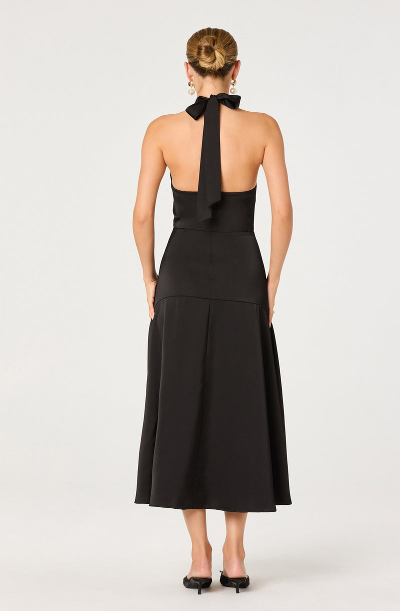 A woman is wearing a sleeveless black dress with a halter neckline tied into a bow at the back. The dress is fitted at the waist and has a full, pleated skirt that falls to a mid-calf length. She is also wearing earrings and black open-toe heels.