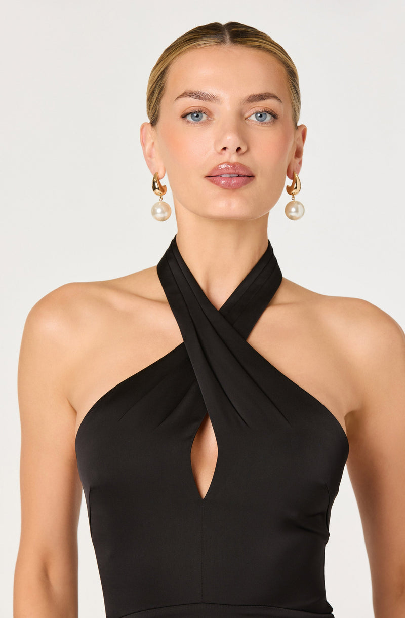 A woman is wearing a black halter dress with a crisscross neckline that creates a keyhole opening in the center. The fabric appears smooth and fitted. She accessorizes with large, dangling pearl earrings with gold accents.