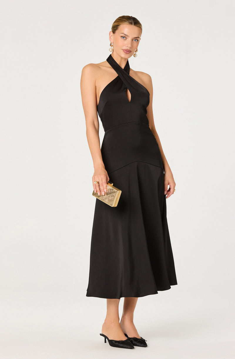 A woman is wearing a sleeveless, black halter-neck dress with a keyhole cutout at the chest. The dress has a fitted bodice and a flared, midi-length skirt. She is holding a small, gold, embellished clutch purse and is wearing black high-heeled shoes with bows on the toes. She has pearl earrings and her hair is styled in a sleek, pulled-back fashion.