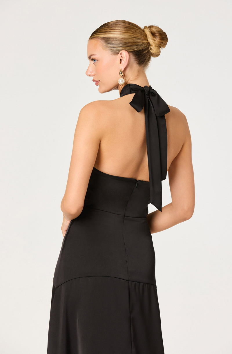 A woman is wearing a black, strapless dress with a fitted bodice and a flowy skirt. The dress features a distinctive bow with long tails tied around the neck in a halter style. She has her hair styled in a sleek, low bun and is accessorized with large, dangling pearl earrings.