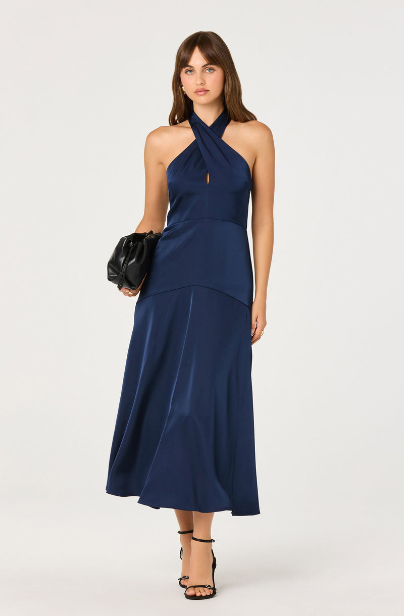 A woman is wearing a navy blue halter-neck maxi dress with a crisscross design at the neckline and a small keyhole cutout in the front. The dress has a fitted bodice and a long, flowing skirt. She is holding a black clutch purse in her right hand and is wearing black strappy high-heeled sandals.