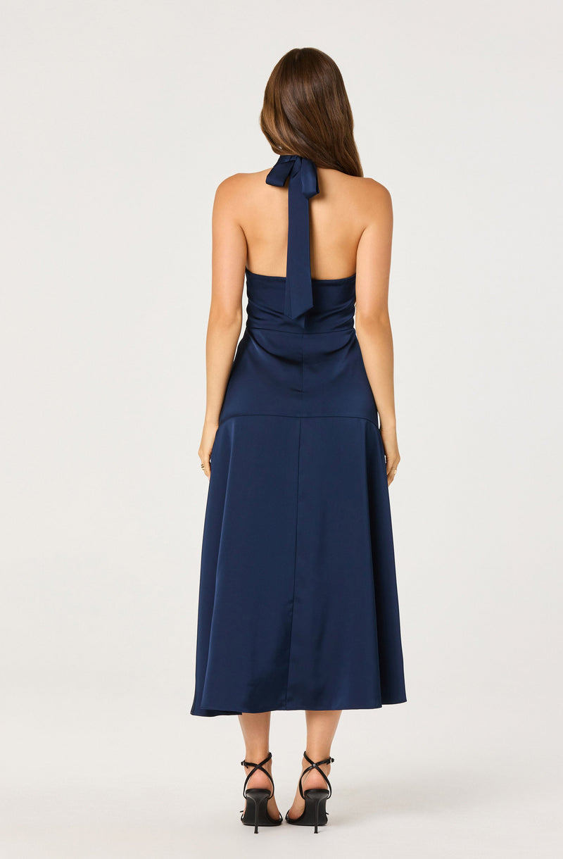 A woman is wearing a strapless navy blue dress with a fitted bodice and a wide waistband. The dress features a halter-style neckline with a large bow at the back of her neck, and the skirt is long, flowing, and reaches mid-calf. She is also wearing black high-heeled sandals with thin ankle straps.