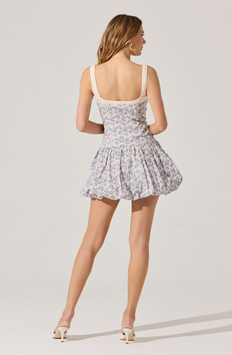 A woman is wearing a sleeveless, fitted dress with wide shoulder straps. The dress has a light-colored background with a delicate blue floral pattern, a square neckline, and a cinched waist. The skirt part of the dress is short, has a gathered, voluminous style, and ends above the knees. She is also wearing white heeled shoes.