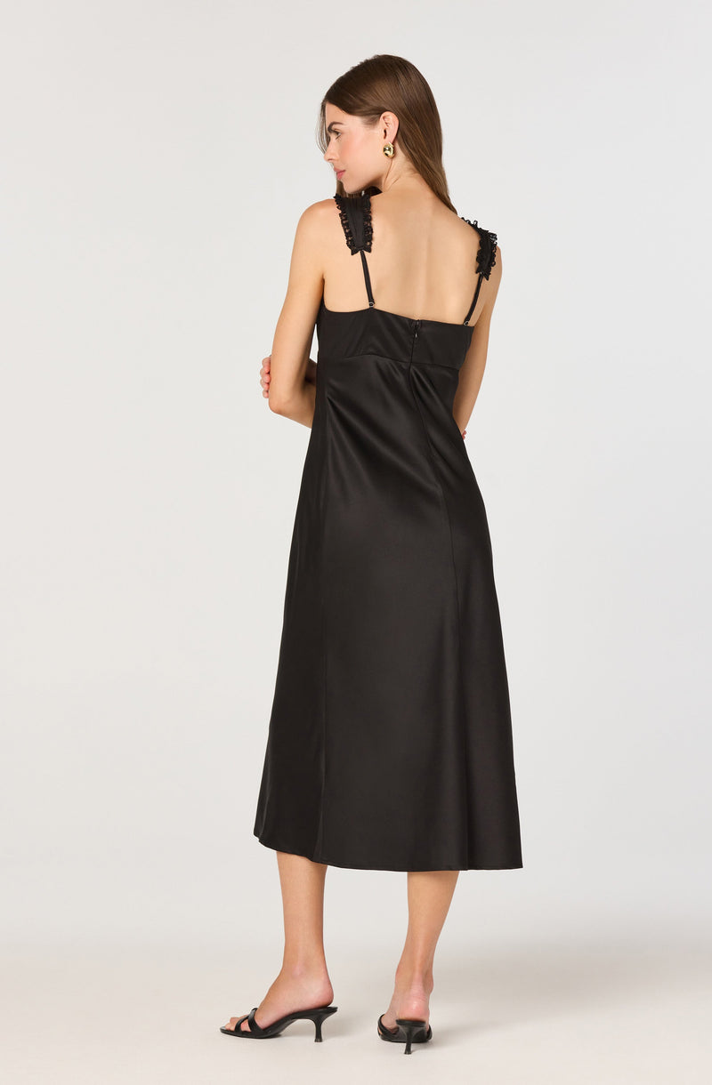 A woman is wearing a black satin sleeveless dress with lace and ribbon details on the shoulder straps. The dress has a fitted bodice and flows down to a midi length. She is also wearing black open-toe heeled sandals.
