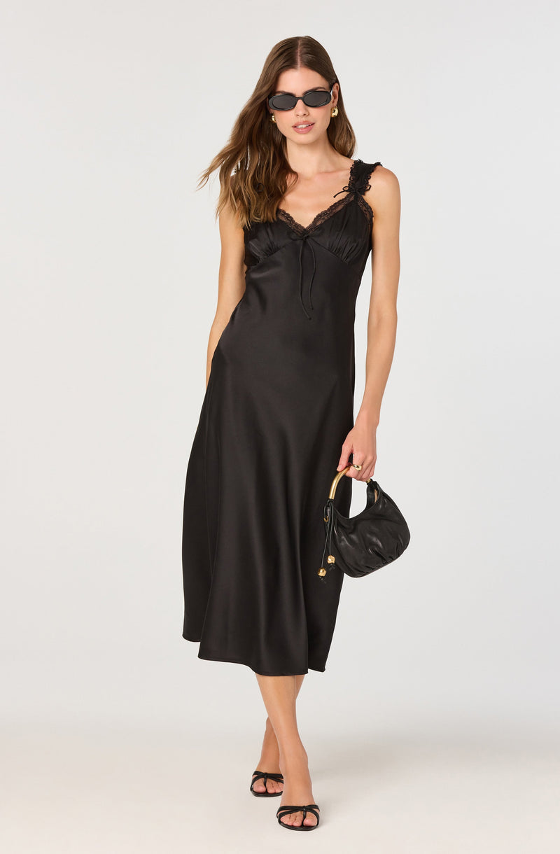 A woman is wearing a black satin slip dress with lace detailing on the straps and neckline. She is accessorizing with black sunglasses, gold hoop earrings, and is holding a black handbag with a gold handle. She is also wearing black open-toe heels.