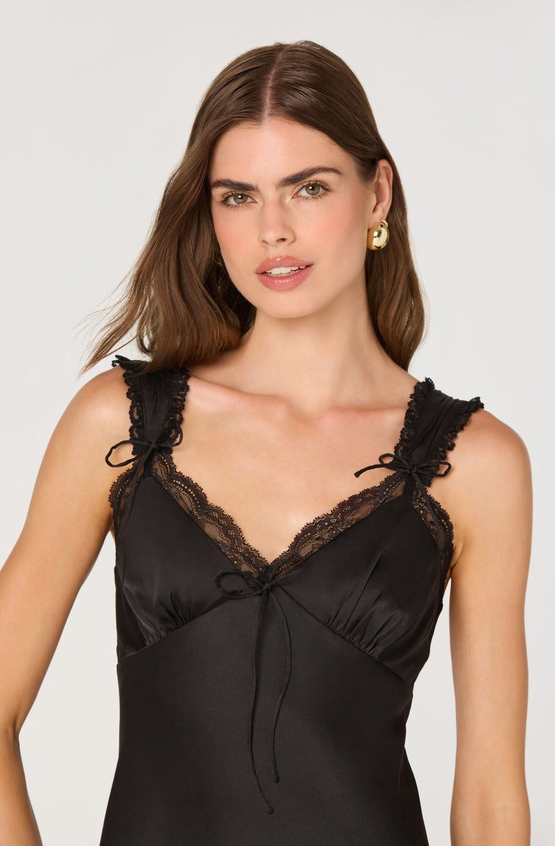 A young woman is wearing a black satin slip dress with lace trim along the neckline and shoulder straps. The shoulder straps are ruffled and tied with small bows. The dress has a fitted bodice and a slight V-neckline. The woman has long brown hair and is wearing a gold earring.