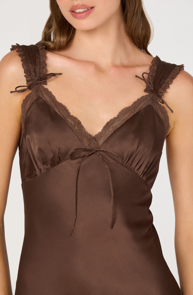 A woman is wearing a satin brown slip dress with lace trim along the V-neckline and shoulder straps. The shoulder straps have small bows, and there is a tied bow at the center of the bust. The dress has a smooth, shiny texture.
