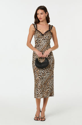 A woman is wearing a sleeveless, leopard print midi dress with black lace detailing on the shoulder straps and neckline. She is holding a small black handbag with a rounded shape and short handle, and is wearing black strappy high-heeled sandals.