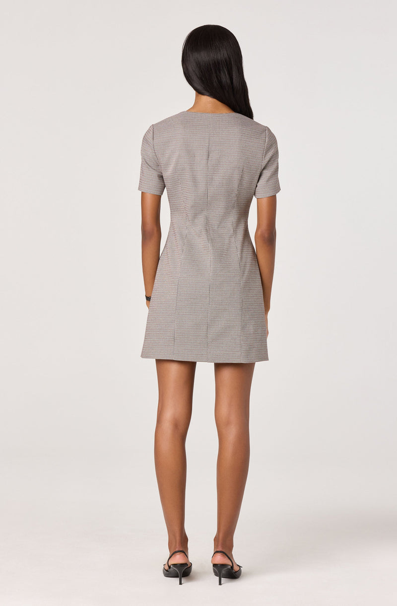 The dress is a short, fitted, plaid pattern with a muted color palette. It has short sleeves with a slight puff at the shoulders and a round neckline. The dress features a subtle checkered design and falls above the knees. The back view shows the dress's tailored fit and clean lines. The woman is wearing black high-heeled sandals.