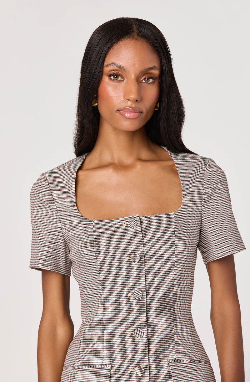 A woman with straight, dark hair is wearing a fitted dress with short sleeves, featuring a small checkered pattern in shades of black, white, and brown. The dress has a square neckline and a row of buttons down the front.