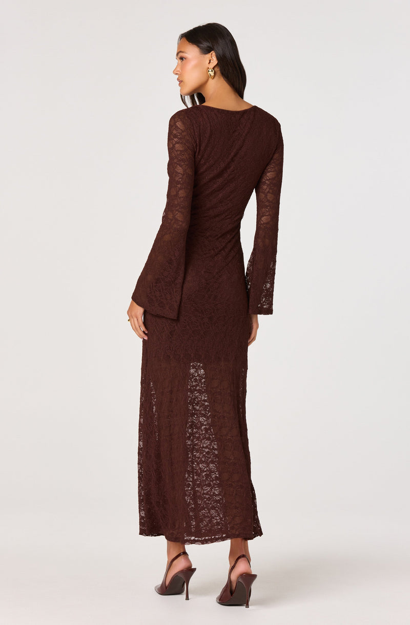 A woman is wearing a long, brown lace dress with a fitted bodice and long, semi-sheer sleeves. The dress has a textured pattern and reaches down to her ankles. She is also wearing pointed high-heeled shoes and large hoop earrings.