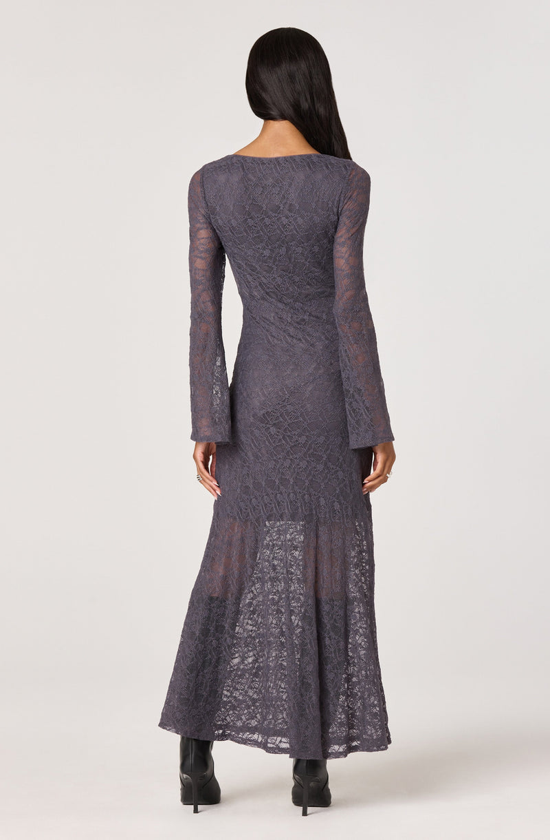 A woman is wearing a long, semi-sheer, dark gray lace dress with intricate patterns. The dress features long sleeves and a fitted bodice, extending down to a flared, slightly transparent skirt that reaches the floor.