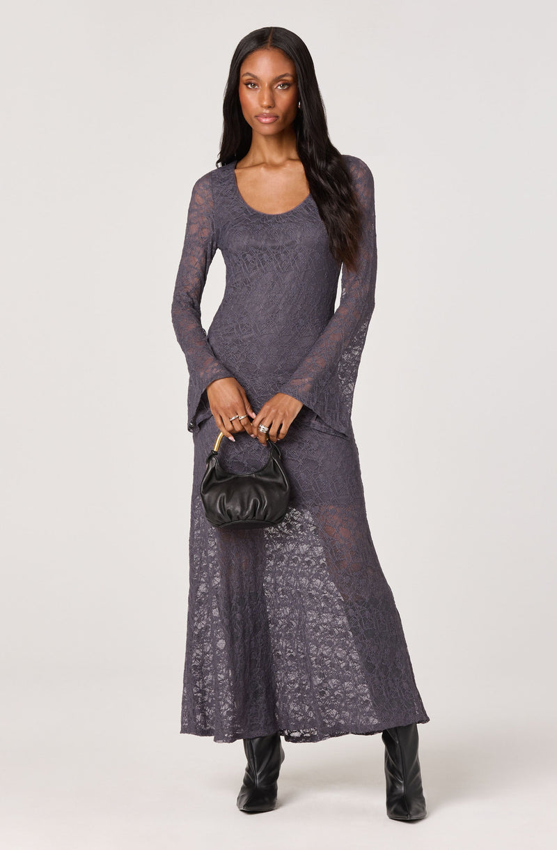 A woman is wearing a matching dark gray lace top and long skirt set that is semi-sheer with a floral pattern. The top has long, slightly flared sleeves and a rounded neckline. She is holding a small black ruched handbag with a gold handle and is wearing black boots.