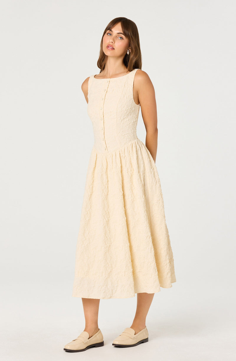A woman is wearing a sleeveless, cream-colored, textured midi dress with a fitted bodice and a full, gathered skirt that falls below the knees. She is pairing the dress with light-colored loafers.