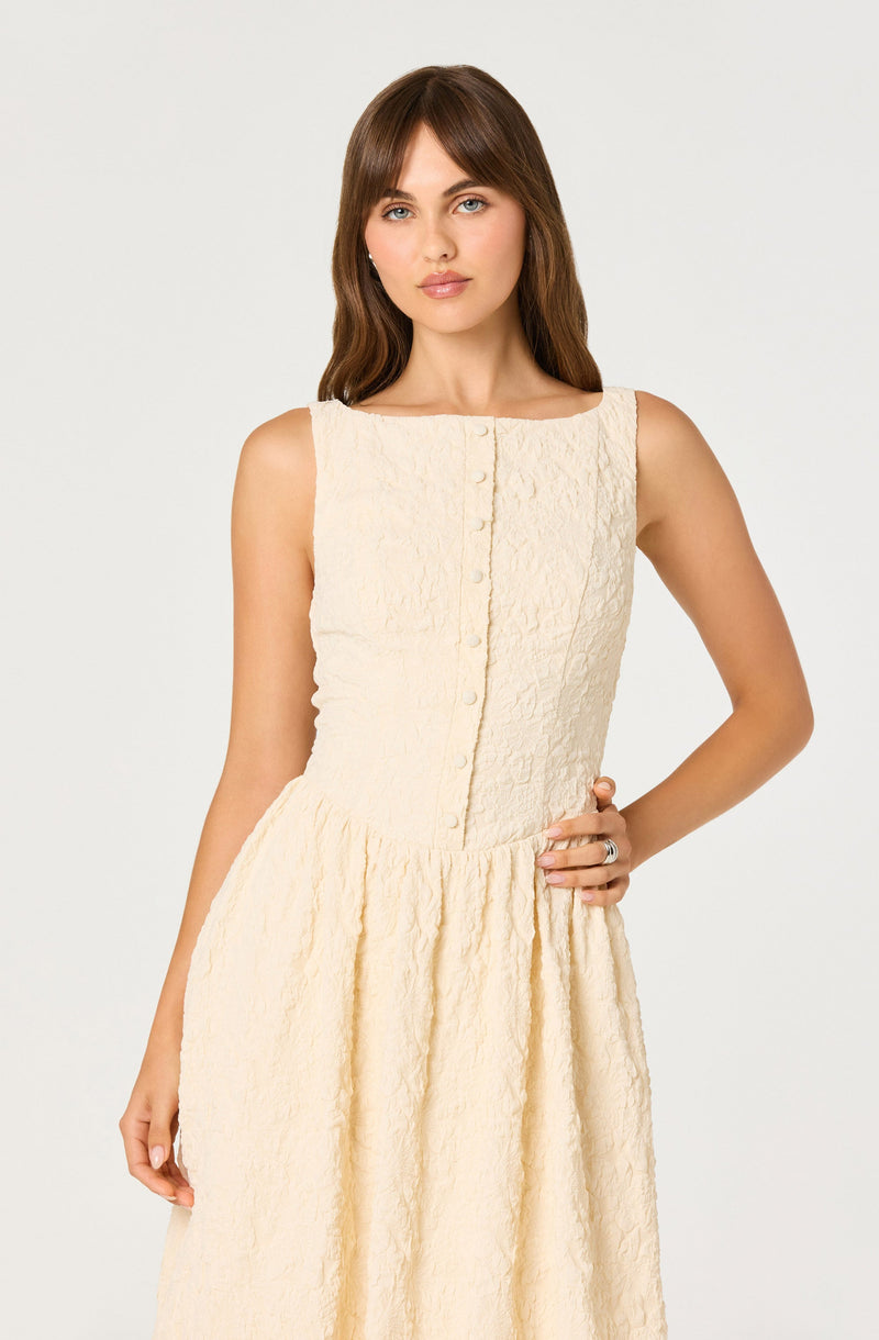 A woman wearing a sleeveless, cream-colored textured dress with a fitted bodice featuring a row of buttons down the front and a full, gathered skirt.