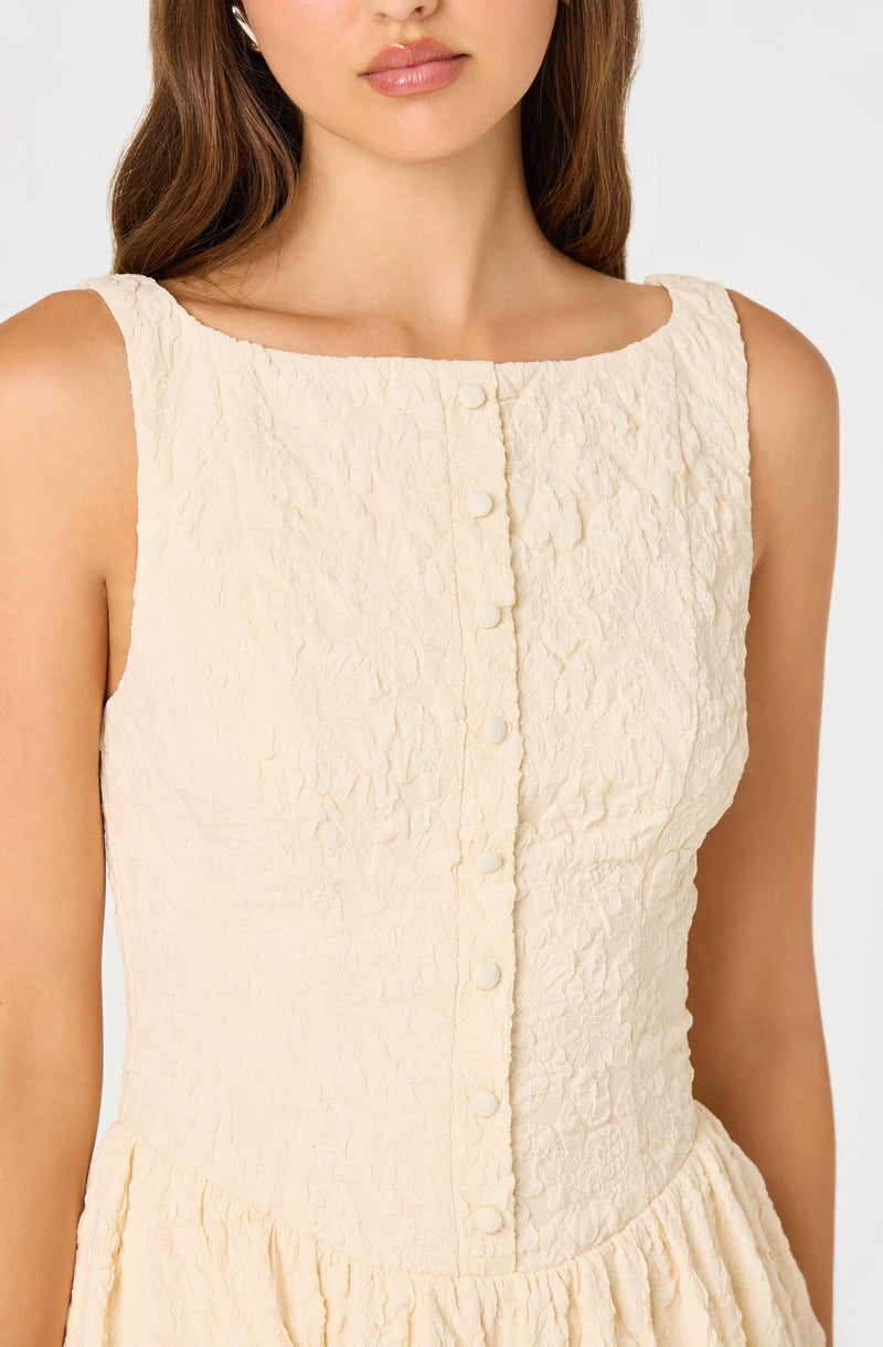 A woman wearing a sleeveless, off-white textured dress with a round neckline. The dress features a row of fabric-covered buttons down the front and a gathered waistline. The fabric has a crinkled, slightly wrinkled appearance.