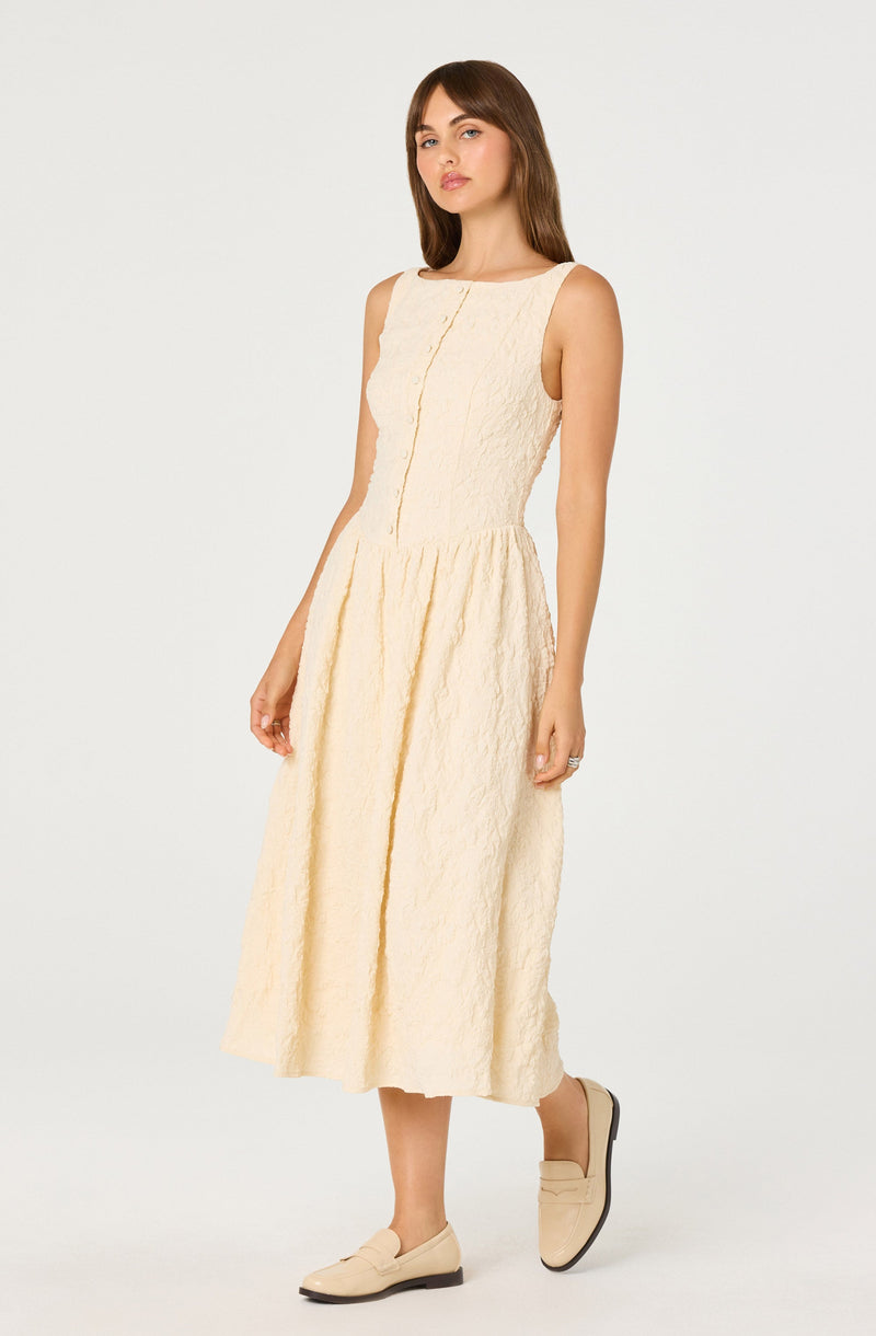 A woman is wearing a sleeveless, cream-colored, textured midi dress with a fitted bodice and a gather at the waist, featuring a front pleat. She has paired the dress with beige slip-on shoes and a ring on her left hand.