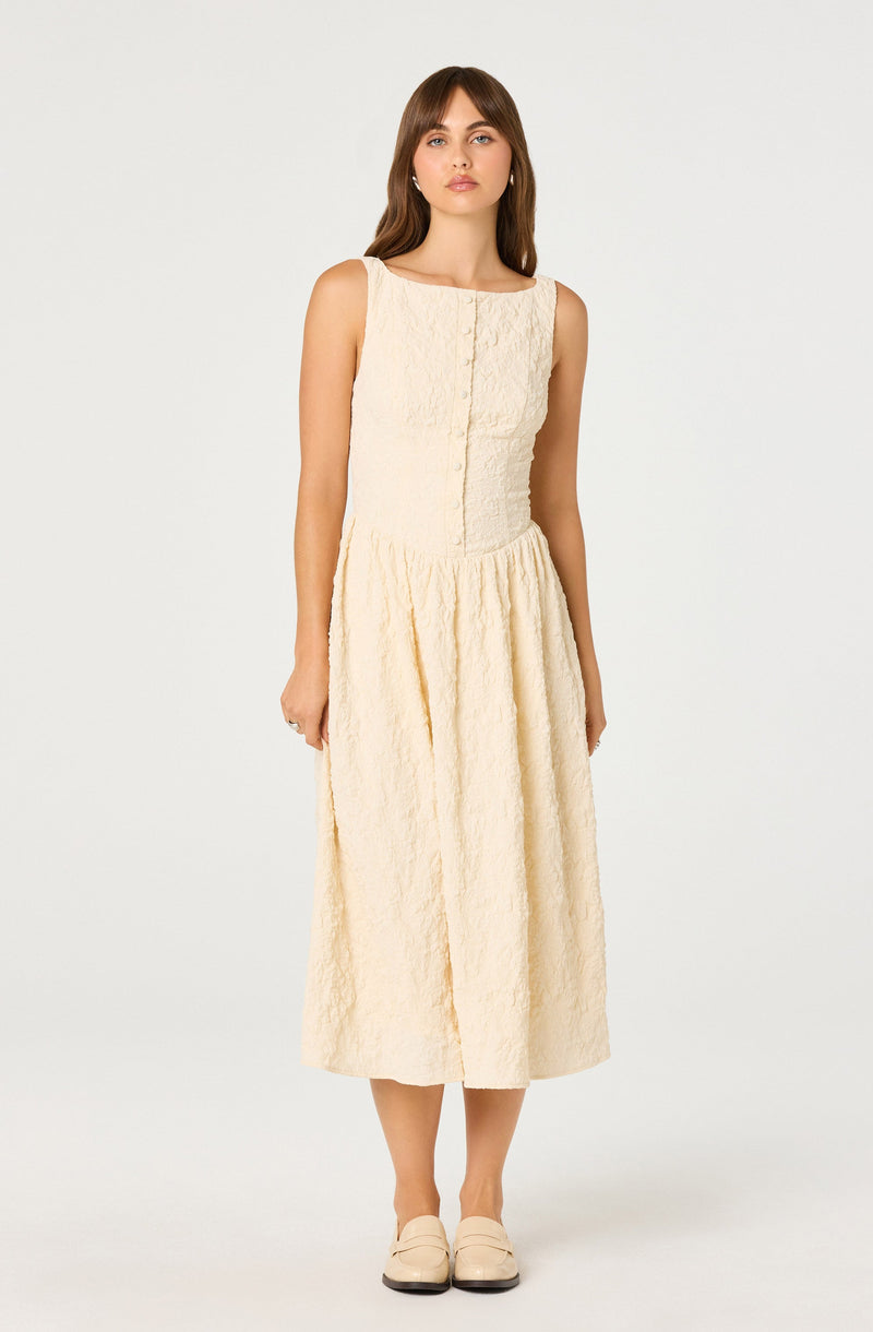 A woman is wearing a sleeveless, cream-colored dress with a textured, slightly crinkled fabric. The dress has a fitted bodice with a subtle vertical seam detail in the center and a gathered, full-length skirt that reaches mid-calf. She is also wearing matching cream-colored shoes.