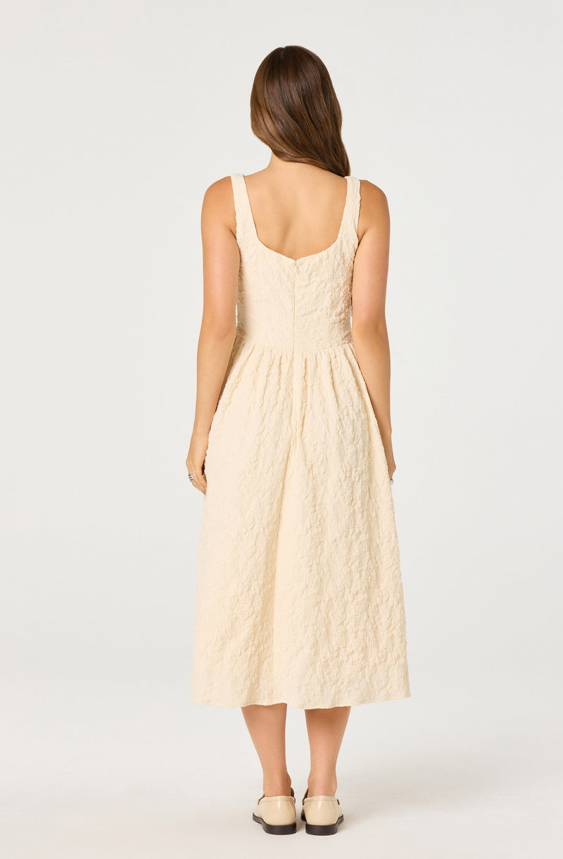 A woman is wearing a sleeveless, ankle-length dress with a fitted bodice and a gathered waist. The dress is made of a textured, cream-colored fabric. She is also wearing beige, low-heeled shoes.