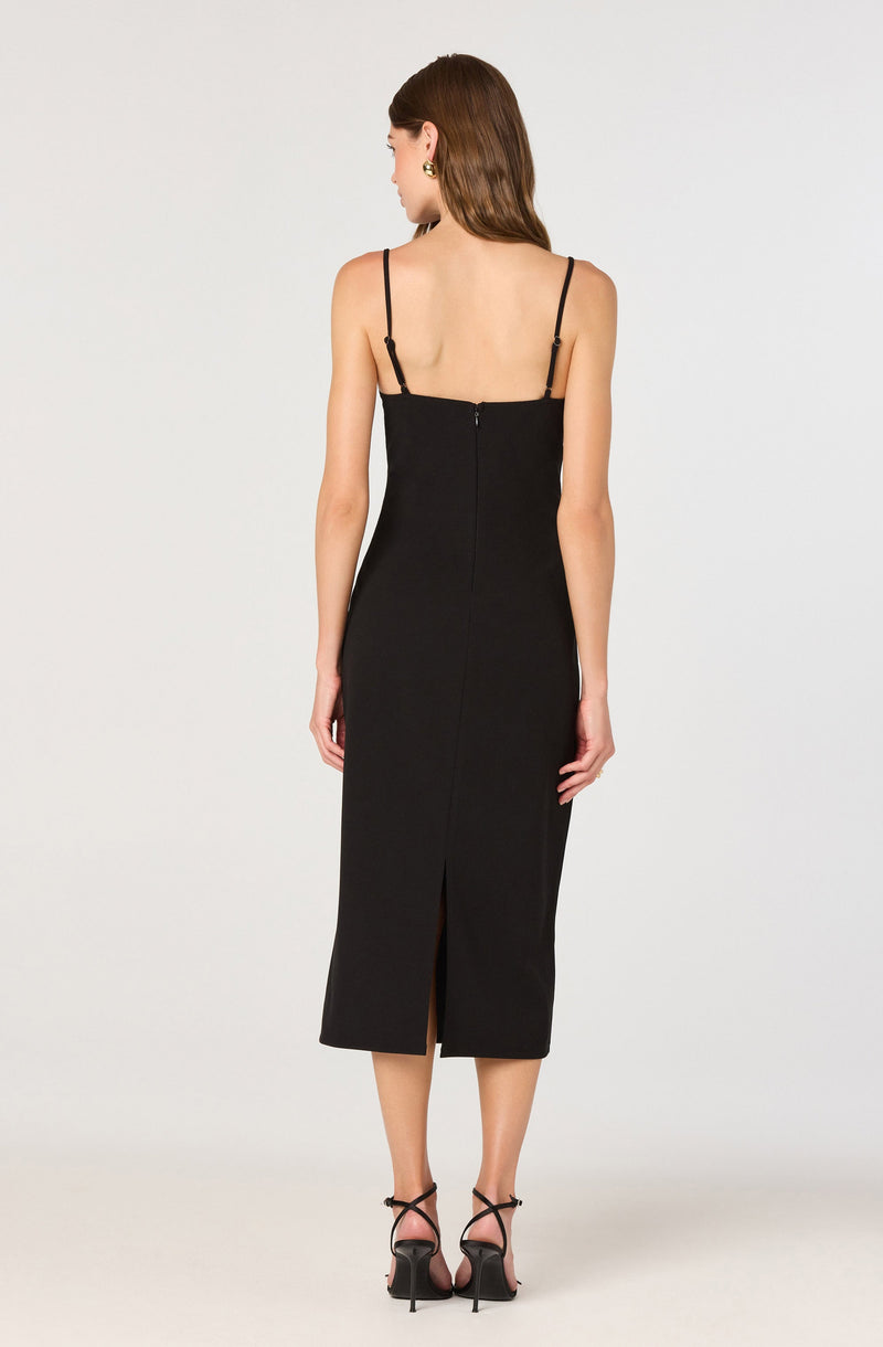 A woman is wearing a long, black sleeveless dress with thin adjustable spaghetti straps. The dress features a back zipper closure and a slit at the center bottom hem. She is also wearing black high-heeled sandals with ankle straps.
