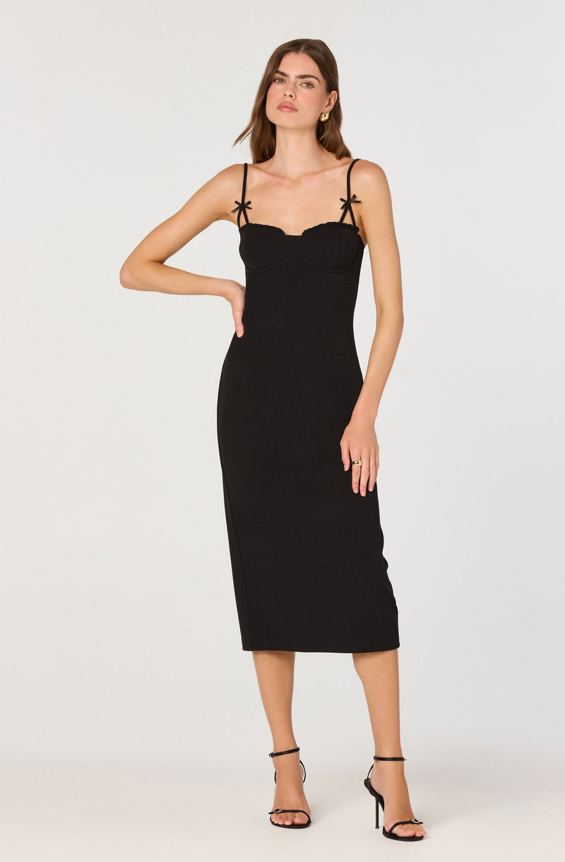 A woman is wearing a black sleeveless dress with thin, tied shoulder straps and a fitted silhouette. The dress has a structured bodice with subtle embellishments. She is also wearing black high-heeled sandals with ankle straps.