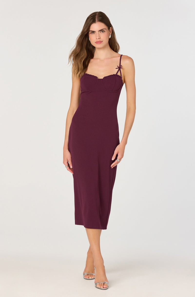 A woman is wearing a sleeveless, fitted, ankle-length dress in a deep burgundy color. The dress features thin spaghetti straps with small bow details on the left side and a sweetheart neckline with lace trim. She is also wearing open-toe high heels with thin straps and subtle jewelry.