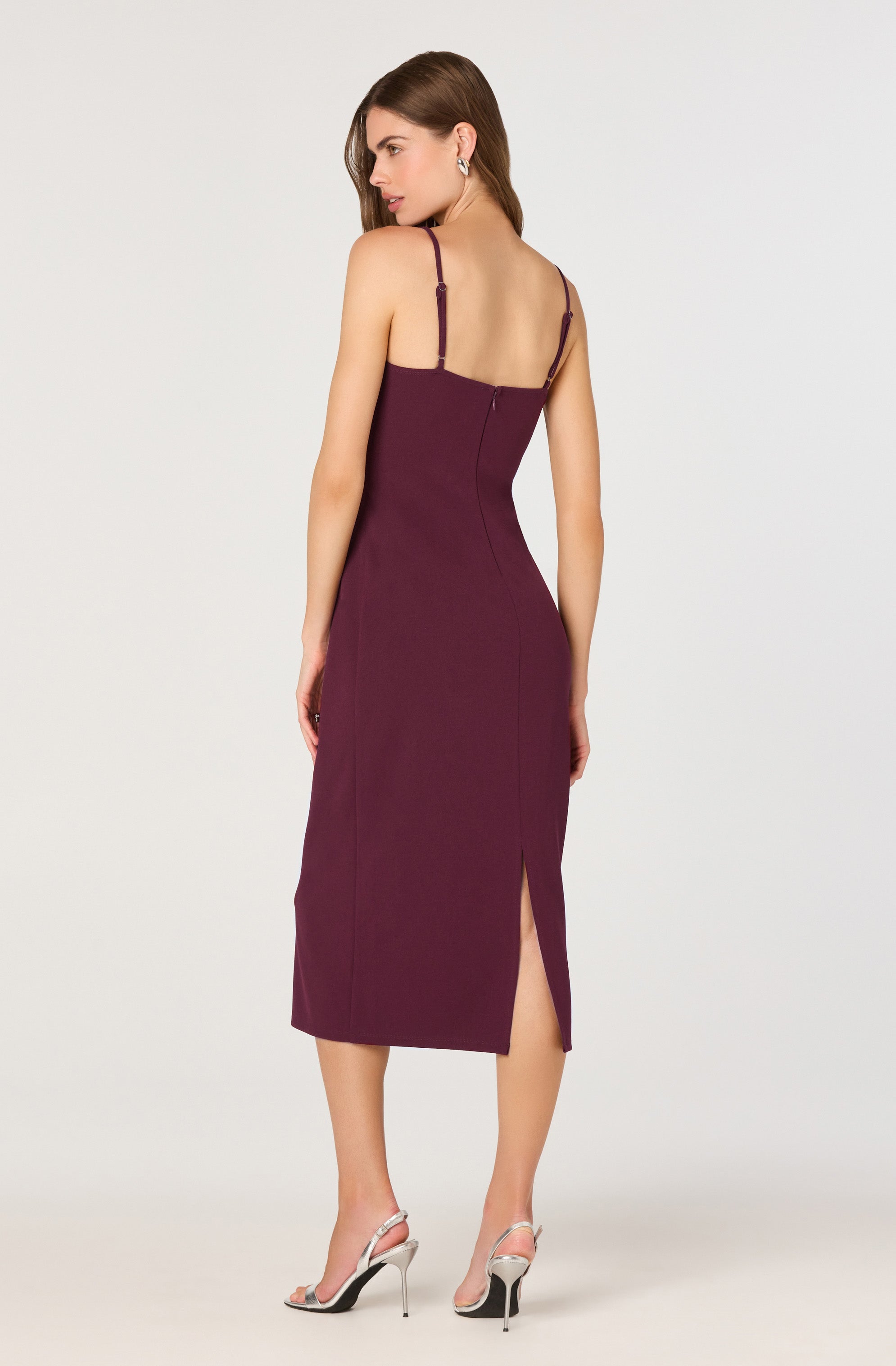 The dress is a sleeveless, fitted, midi-length garment in a deep burgundy color. It features thin adjustable shoulder straps, a back zipper closure, and a front slit that extends to just below the knee. The fabric appears smooth and structured, accentuating the waist and hips. The model pairs the dress with high-heeled sandals that have a thin ankle strap and stiletto heel.