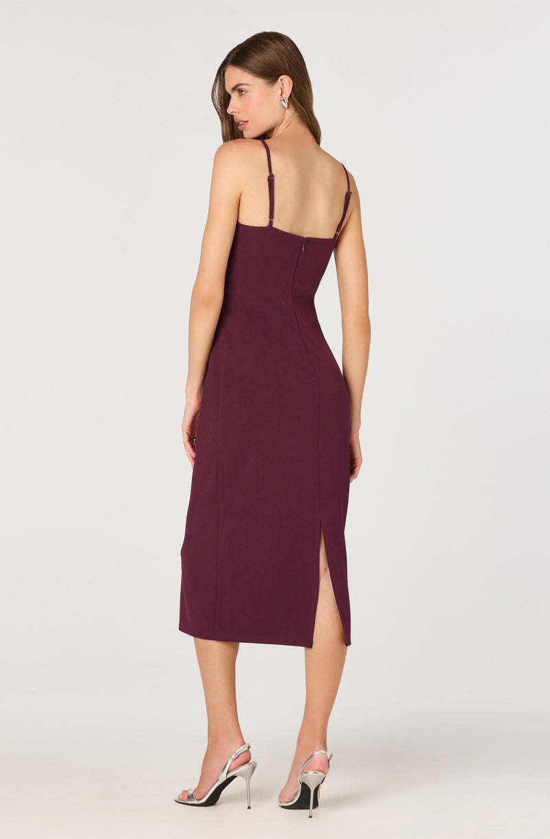 The dress is a sleeveless, fitted, midi-length garment in a deep burgundy color. It features thin adjustable shoulder straps, a back zipper closure, and a front slit that extends to just below the knee. The fabric appears smooth and structured, accentuating the waist and hips. The model pairs the dress with high-heeled sandals that have a thin ankle strap and stiletto heel.