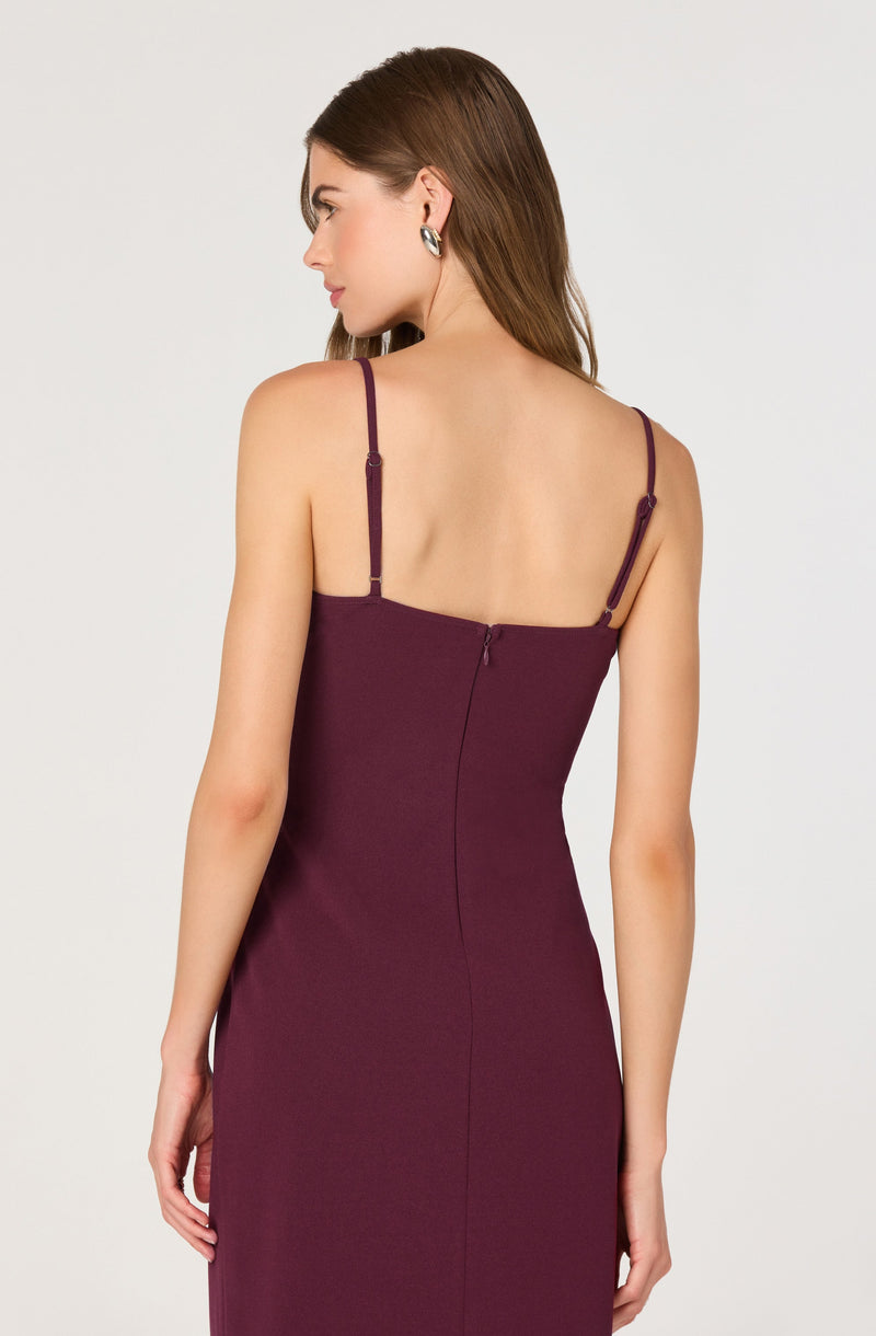 A woman is wearing a fitted, sleeveless dress in a deep burgundy color with thin, adjustable spaghetti straps. The back of the dress features a concealed zipper closure.