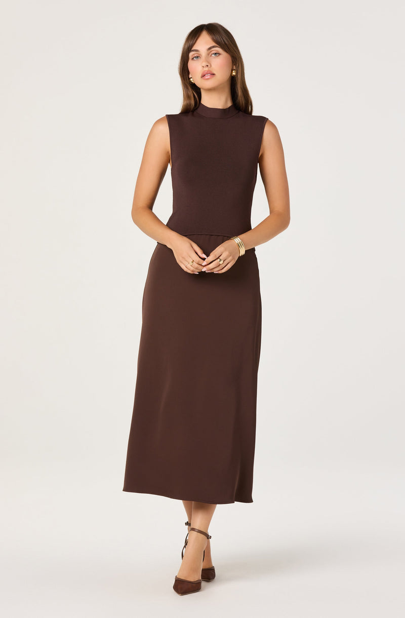 A woman is wearing a sleeveless, high-neck, dark brown top paired with a long, matching dark brown skirt that reaches her ankles. She has accessorized with gold hoop earrings, a gold bracelet, and rings, and is wearing brown high-heeled shoes with ankle straps.