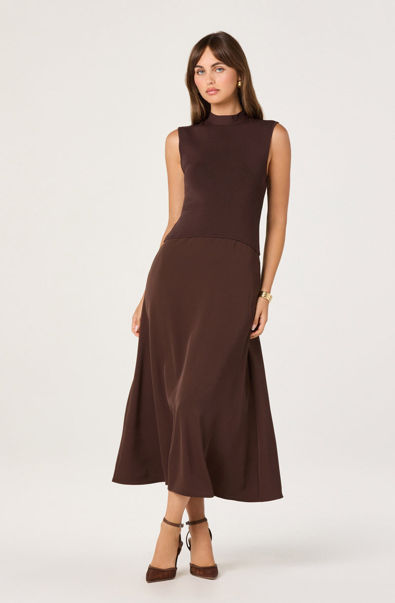 The woman is wearing a sleeveless, high-neck, dark brown top paired with a long, flowing, slightly shiny brown skirt. She has on brown ankle-strap heels and gold accessories, including earrings and a bracelet.