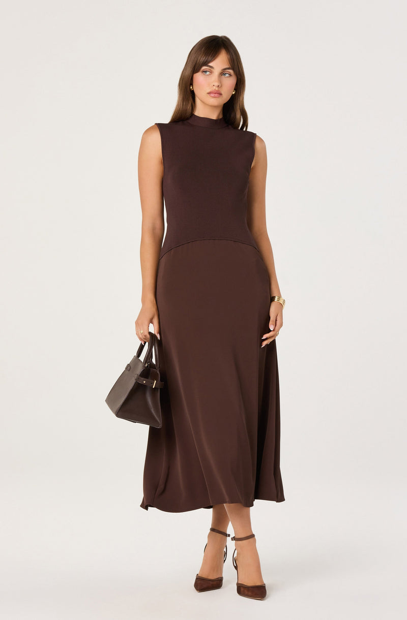 A woman is wearing a sleeveless, high-neck, ankle-length brown dress with a fitted bodice and a slightly flared skirt. She is holding a medium-sized brown handbag in her left hand and is wearing matching brown high-heeled sandals. She has small gold earrings and a gold bracelet on her right wrist.