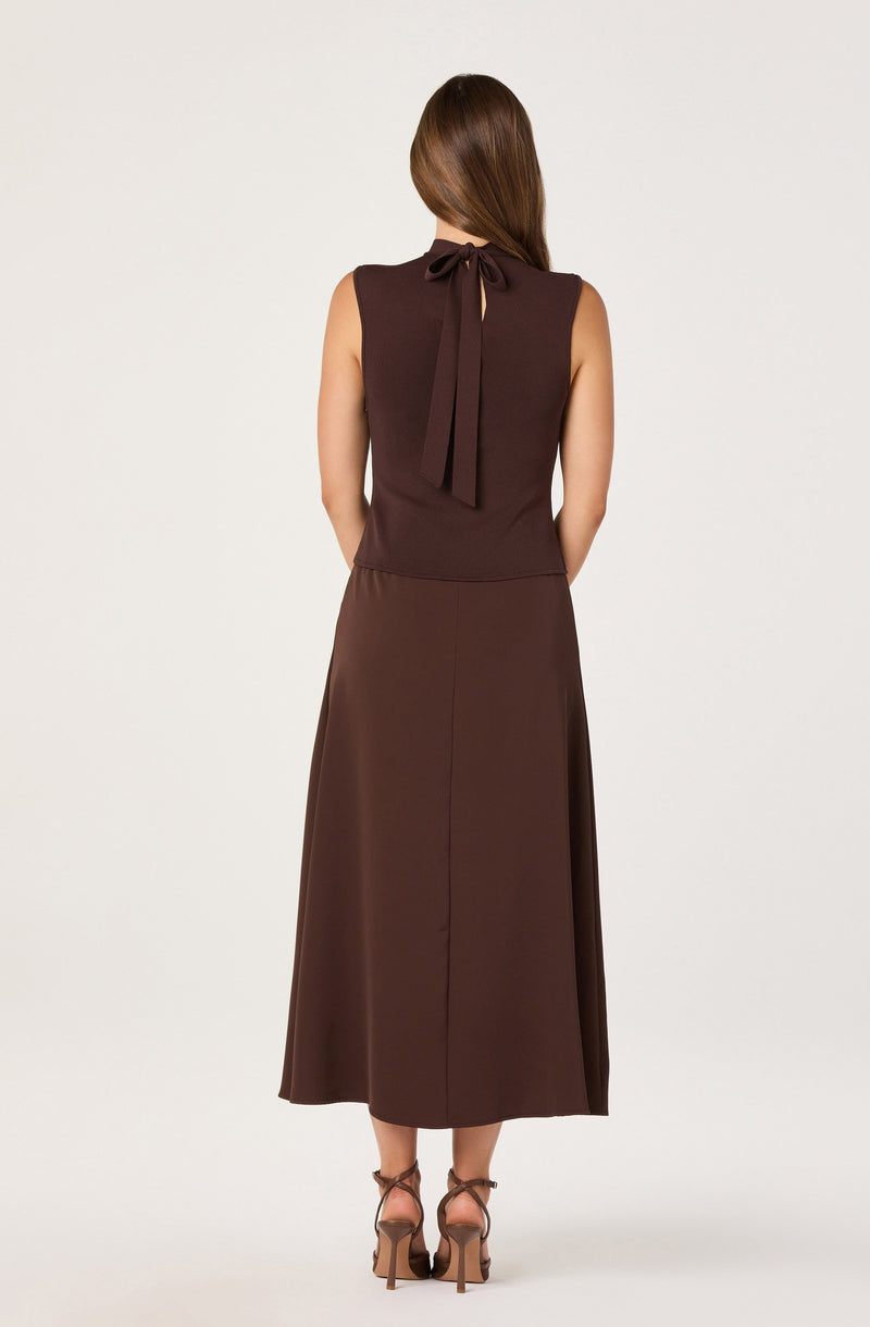 The woman is wearing a sleeveless, dark brown dress with a fitted waist and a full, flowing skirt that reaches mid-calf. The dress features a bow-tie detail at the back neckline. She is also wearing a pair of high-heeled, strappy sandals.