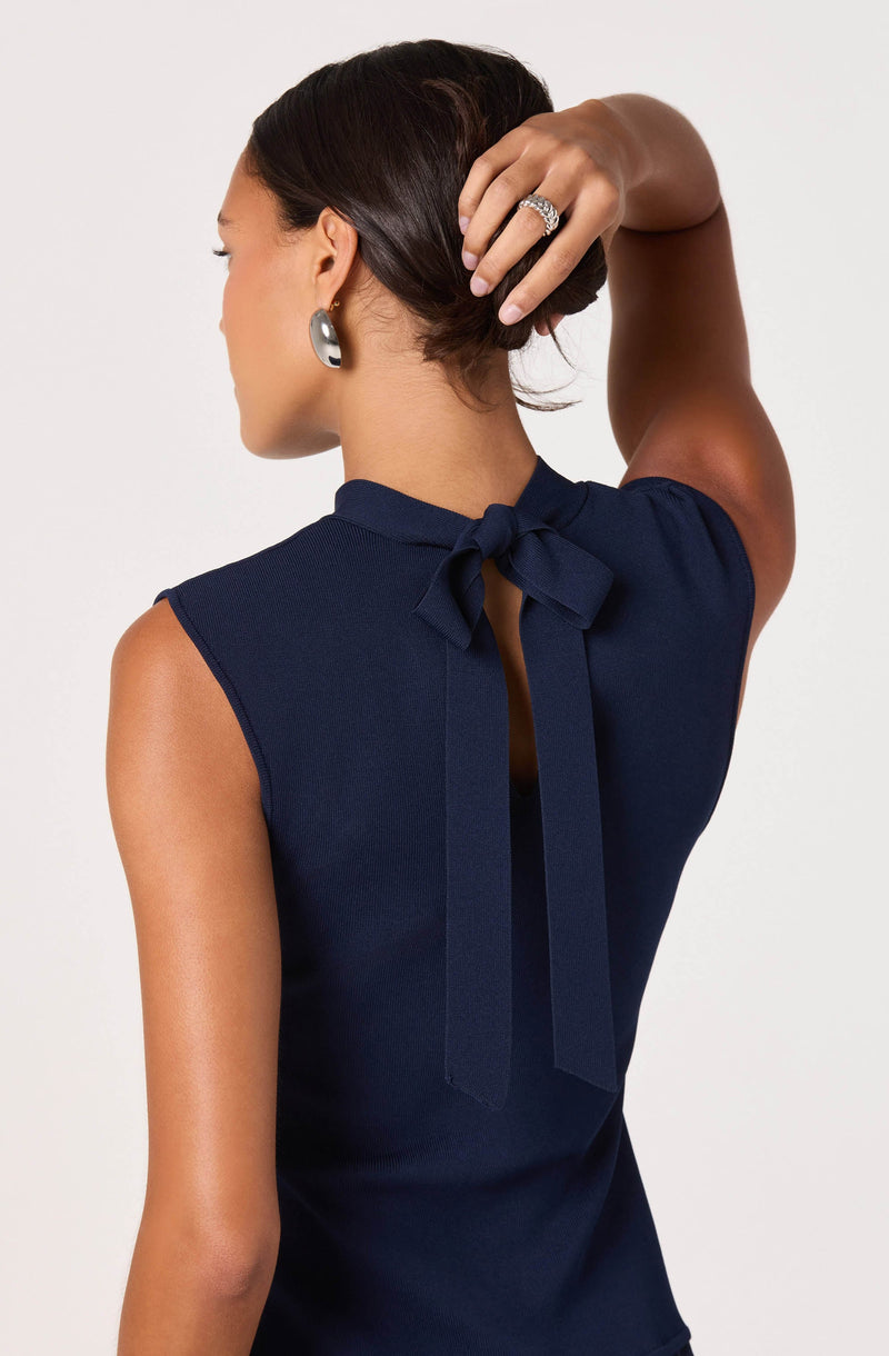 A woman is wearing a sleeveless navy blue top with a bow tied at the back of the neckline. The top has a small keyhole opening below the bow. She accessorizes with silver hoop earrings and a large ring on her right hand.