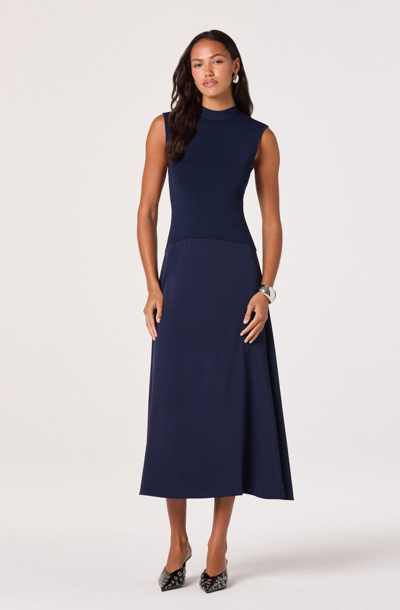 The woman is wearing a sleeveless, navy blue, high-neck dress that is fitted at the top and flares out into a long, flowing skirt. She has on patterned high heels and a wristwatch, with her hair styled in loose waves.