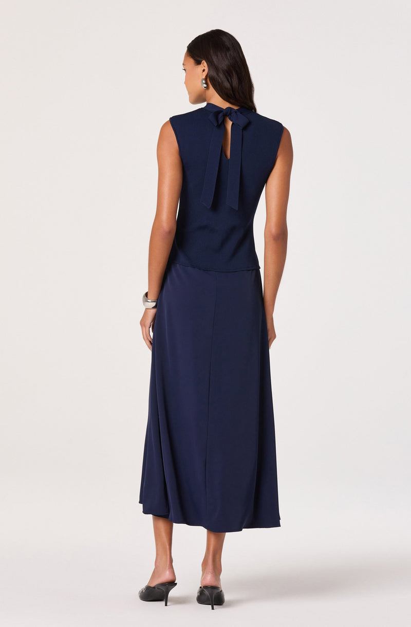 A woman is wearing a sleeveless navy blue dress with a fitted bodice and a long, flowing skirt. The dress features a keyhole opening at the back, tied with a large bow at the neckline. She accessorizes with pearl earrings and a wide bracelet on her right wrist. She is also wearing black high-heeled shoes.