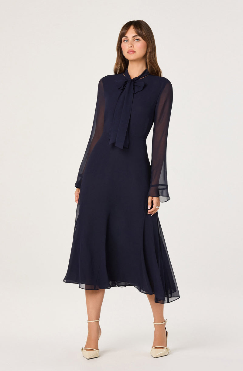 A woman is wearing a navy blue dress with sheer, long sleeves and a bow-tie collar. The dress is midi-length and flowy, with a slightly asymmetrical hemline. She is also wearing beige high-heeled shoes with ankle straps.