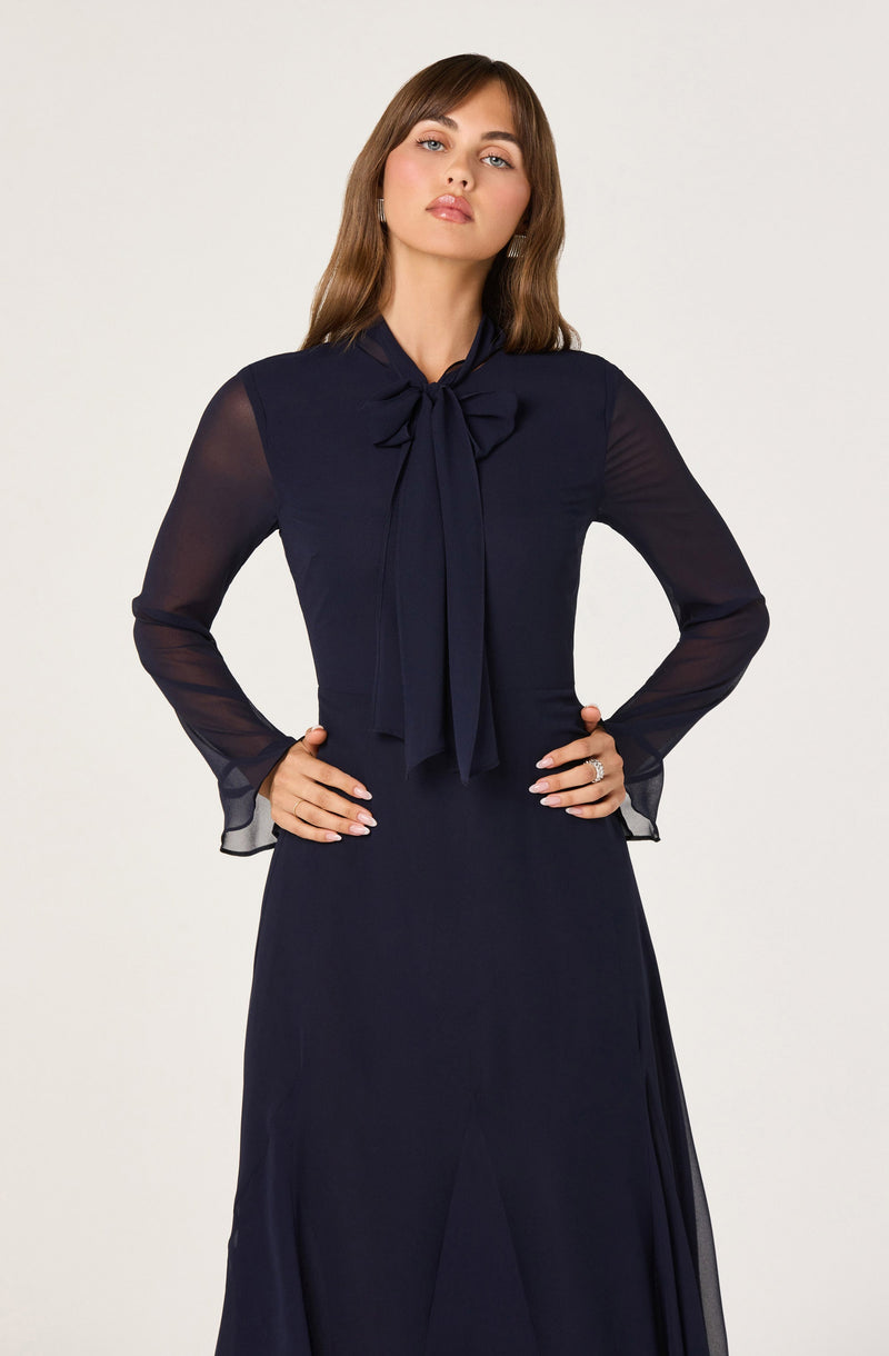A woman is wearing a long, navy blue dress with sheer, long sleeves. The dress features a high neck with a large bow tied at the neck.