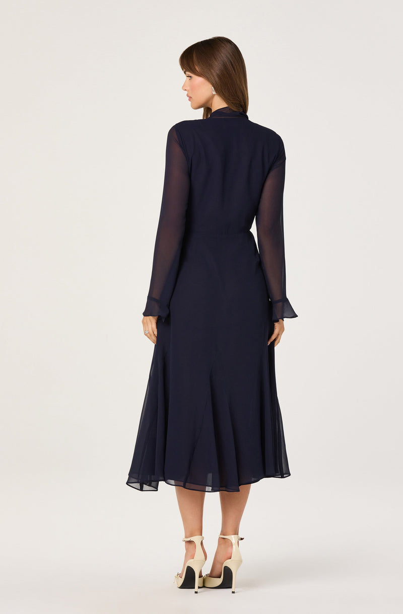 A woman is wearing a long, navy blue dress with sheer, long sleeves and a fitted bodice. The skirt is flowing and reaches mid-calf length. She pairs the dress with beige high-heeled sandals featuring ankle straps.