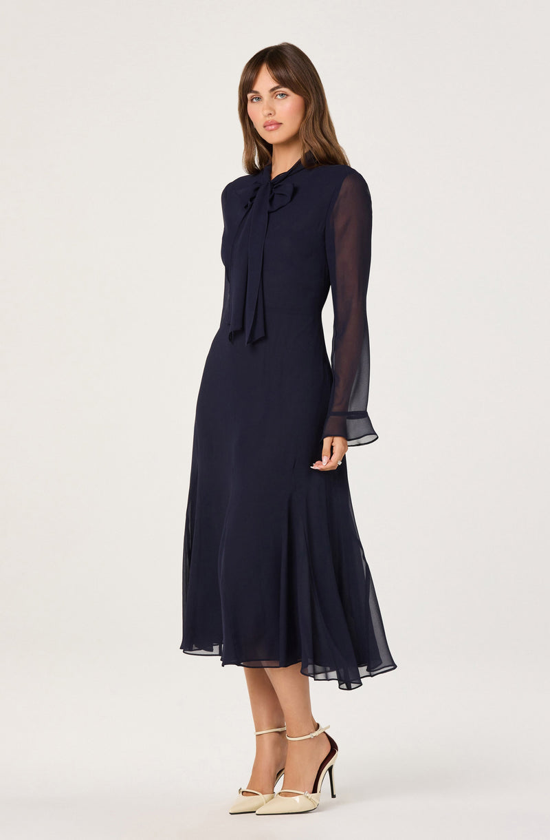 The woman is wearing a long, navy blue dress made of a sheer, flowing fabric. The dress features a high neckline with a large bow tie at the collar. The long sleeves are semi-transparent, with a slight puff at the shoulders and a flared, sheer cuff at the wrists.