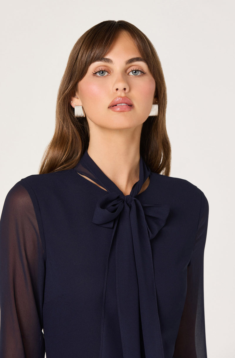 A woman is wearing a navy blue blouse with a large bow tied at the collar. The blouse has sheer long sleeves and a small cut-out detail near the neckline.