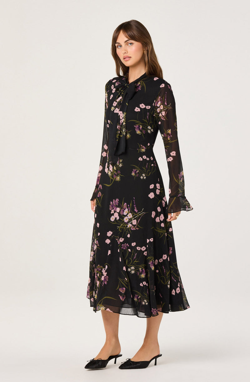 A woman is wearing a long, black, semi-sheer dress with a floral pattern in shades of pink, purple, and green. The dress features a fitted waist, long sleeves with ruffled cuffs, and a midi-length hem. She has a black bow-tie scarf around her neck and is wearing black pointed-toe heels with a small bow detail.