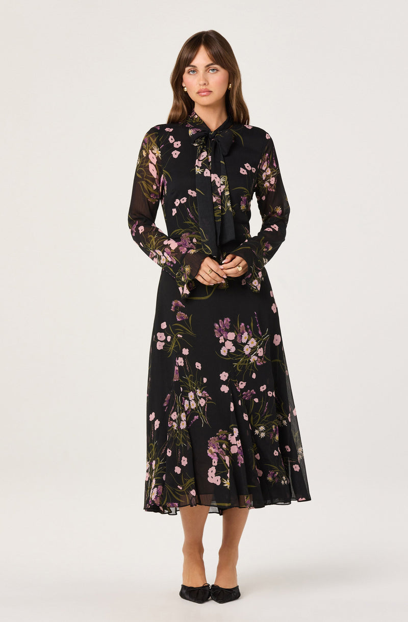A woman is wearing a long, black dress with a floral pattern featuring pink, purple, and green hues. The dress has a sheer, lightweight fabric with a flowy silhouette and a midi length. She pairs the dress with a matching sheer blouse that has a large bow at the neckline, long sleeves, and a similar floral print. She completes her outfit with black pointed-toe shoes that have a bow detail.