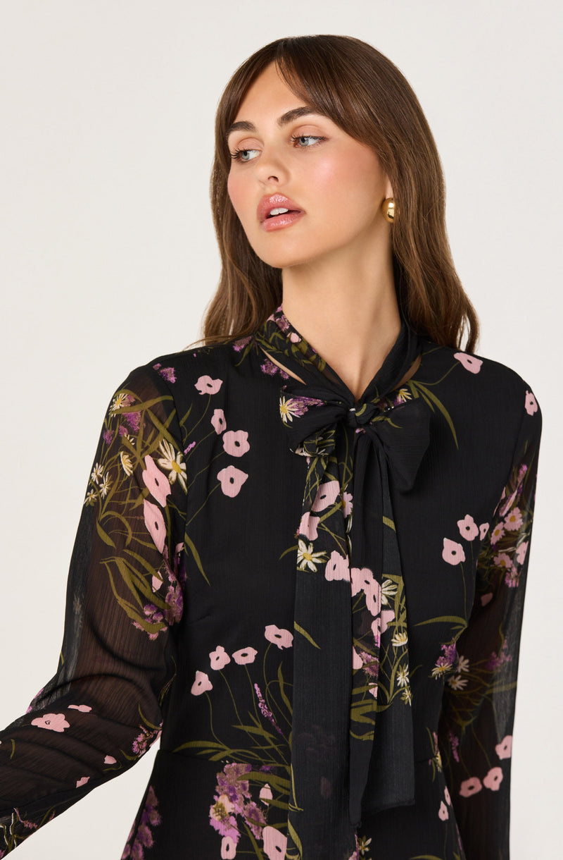 A woman is wearing a black sheer blouse with a floral pattern featuring pink, purple, and white flowers with green leaves. The blouse has a high neckline with a large bow tied at the front.