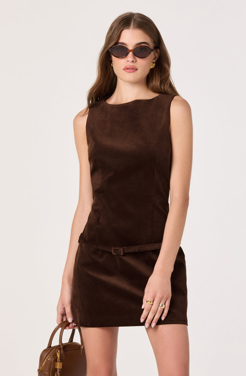A woman is wearing a sleeveless, dark brown suede dress with a round neckline and a small matching belt at the waist. She accessorizes with black oval sunglasses, gold hoop earrings, and gold rings on her fingers. She is holding a small brown handbag in her left hand.