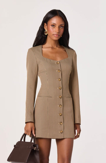 A woman is wearing a fitted, knee-length dress made of a light brown fabric with a small checkered pattern. The dress has long sleeves, a square neckline, and features a row of large, gold buttons down the front. She is holding a dark brown handbag in her right hand and has large, gold hoop earrings.