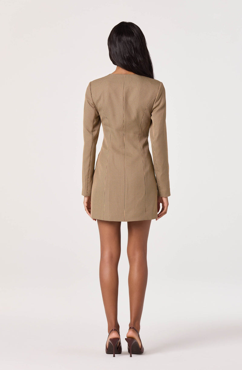 The woman is wearing a fitted, long-sleeve, camel-colored mini dress with a subtle pinstripe pattern. She is also wearing a pair of high-heeled shoes.