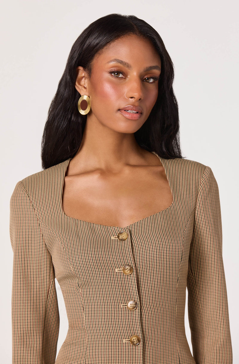 A woman is wearing a fitted, long-sleeved blazer with a small checkered pattern in shades of beige, brown, and black. The blazer features a square neckline and is fastened with large, shiny gold buttons down the front. She also has large, gold hoop earrings.