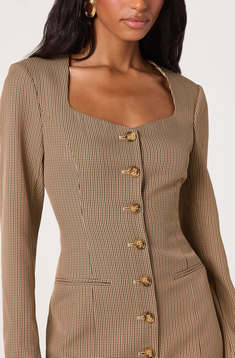 A woman is wearing a fitted blazer with a small checkered pattern in shades of brown, black, and beige. The blazer features a square neckline and is fastened with a row of large, round, gold buttons down the front. She is accessorized with large, gold hoop earrings.